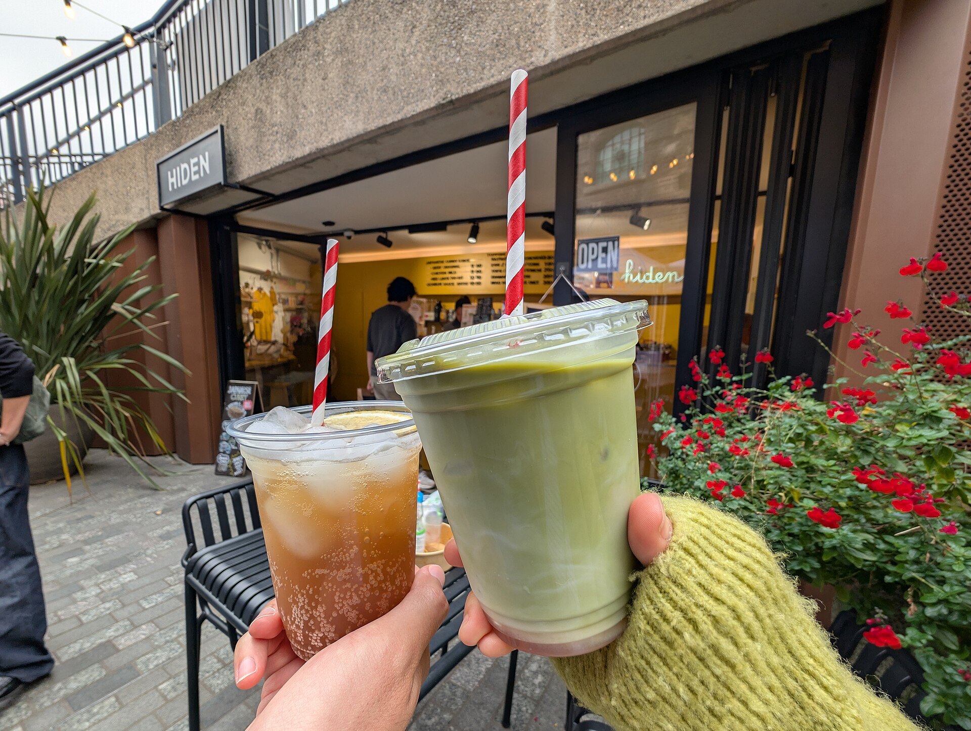Ein Glas Iced Sakura Matcha Latte wird vor einem Café in die Kamera gehalten.